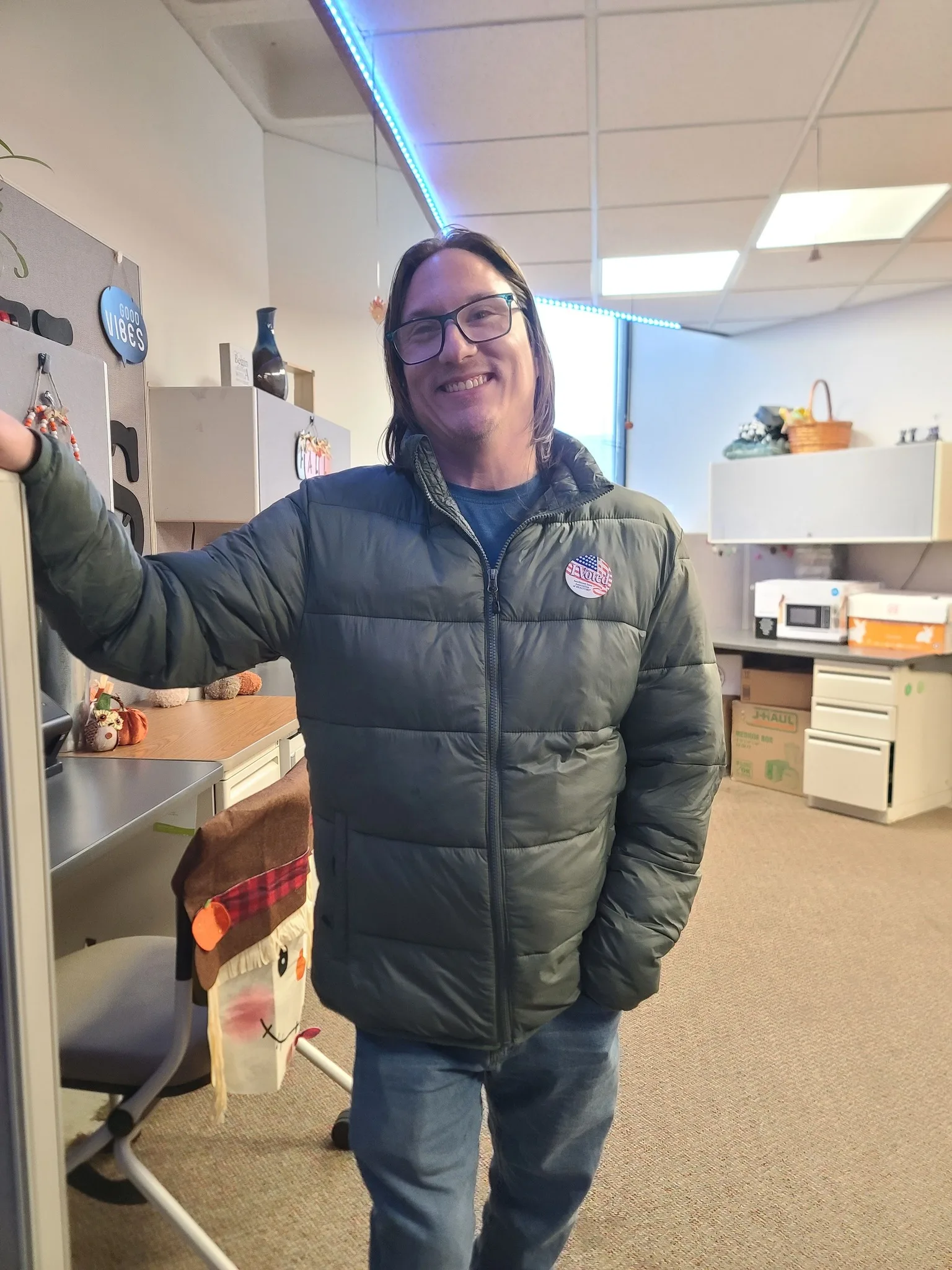 Tim stands smiling in an office setting, proudly wearing his "I Voted" sticker after participating in the election.